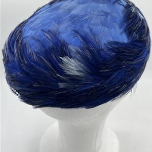 May include: A vintage blue feather hat, displayed on a white mannequin head. The hat features a mix of dark and light blue feathers, creating a textured and layered effect. The feathers are arranged to cover the entire hat.