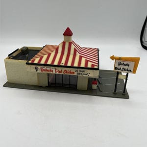 May include: Miniature Kentucky Fried Chicken restaurant model with a red and white striped roof and a sign that reads "Kentucky Fried Chicken." The building is beige with a red trim and a small parking lot.