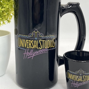 Puede incluir: Juego de taza y vaso de chupito negros de Universal Studios Hollywood. La taza tiene un asa grande y las palabras "UNIVERSAL STUDIOS Hollywood" en amarillo, azul y morado. El vaso de chupito tiene el mismo logotipo.