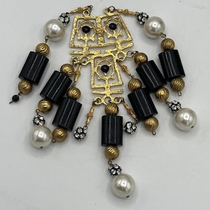 Puede incluir: Un collar dorado con un diseño geométrico. El collar presenta cuentas cilíndricas negras, perlas de imitación y cuentas doradas y con cristales más pequeñas. El diseño central tiene formas caladas con detalles en negro.