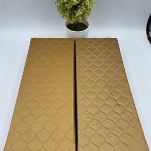 May include: Two rectangular, gold-colored cushions with a quilted, diamond-patterned design. The cushions are placed side-by-side, and a small, artificial plant in a white pot sits above them.
