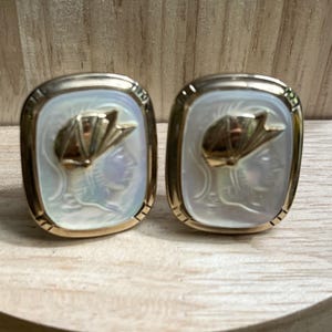 May include: A pair of gold-toned cufflinks with a rectangular shape. Each cufflink features a white, iridescent background with a gold-colored relief of a classical figure. The cufflinks are displayed on a light-colored wooden surface.