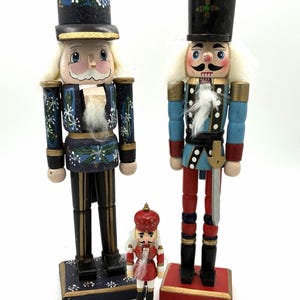 Puede incluir: Tres figuras de cascanueces de madera de pie juntas. El cascanueces de la izquierda es azul oscuro con patrones de copos de nieve blancos. El de la derecha lleva una chaqueta azul claro con pantalones rojos y una espada. Un pequeño cascanueces rojo está en el centro.