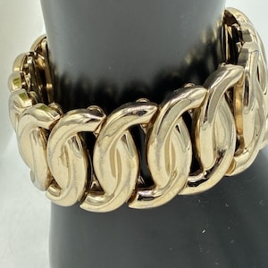 May include: A gold-toned metal bracelet featuring a series of interlocking, curved, and polished links. The bracelet has a substantial, wide design with a reflective surface that catches the light, creating a continuous, repeating pattern around the wrist.