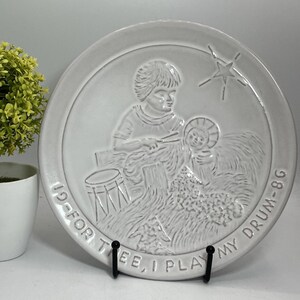 May include: A white ceramic plate with a raised relief design showing a child playing a drum for the baby Jesus. The plate has the text "I PLAY MY DRUM" and "FOR THEE". The plate is displayed on a black stand.