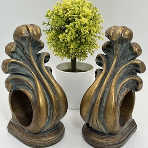 May include: Pair of decorative bronze-colored napkin rings with an ornate, leaf-like design. The rings have a distressed finish with hints of teal and sit on a rounded base. A small, artificial green plant in a white pot is in the background.