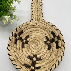 Può includere: Un cestino rotondo intrecciato con manico. Il cestino presenta un colore beige naturale con un motivo geometrico nero al centro. Il bordo e il manico sono rinforzati con cuciture marrone scuro.