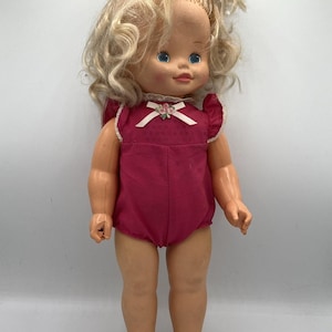 May include: A vintage doll with blonde, curly hair and blue eyes. The doll wears a magenta-colored romper with a white bow and floral detail at the neckline. The doll's arms and legs are slightly bent, and it stands on a white surface.