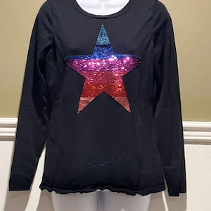 May include: A black long-sleeve shirt featuring a large, centred star graphic made of multi-coloured sequins. The star transitions from a teal top point to a blue middle section, followed by a vibrant pink centre and a red base. The shirt has a round neckline.