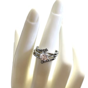 Peut inclure: Une bague argentée dotée d'une pierre centrale ronde et transparente sertie dans un design à plusieurs anneaux. Les deux anneaux extérieurs sont ornés de petites pierres transparentes pavées qui se courbent vers le centre, créant un effet superposé.