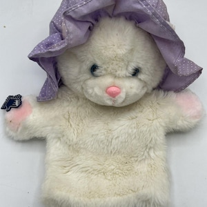 May include: A white plush hand puppet shaped like a cat, wearing a lavender bonnet with white polka dots. The puppet has blue eyes, a pink nose, and pink paws. A small tag is attached to the puppet.