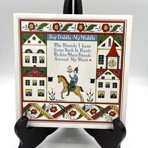 May include: A square ceramic tile featuring a central illustration of a person riding a horse, surrounded by stylized houses and floral borders. The text reads: Hey Diddle My Middle, The Pounds I Lose Come Back In Haste To Join Their Friends Around My Waist.