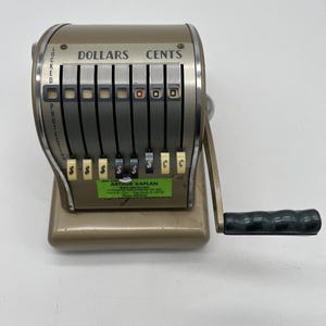 May include: Vintage gold-toned mechanical cash register with a crank handle and multiple dollar and cent indicators. The register has a series of levers and a green label with text. The word "DOLLARS" and "CENTS" are visible on the top.
