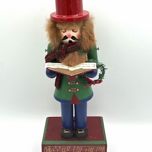May include: A wooden Christmas nutcracker figurine. The figurine wears a red top hat, green coat, blue pants, and a burgundy scarf. It holds a book with musical notes and a small wreath. The base is red with musical notes.
