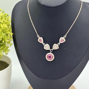 May include: A silver-toned necklace featuring a central circular pendant with a bright pink faceted stone surrounded by a halo of clear crystals. The chain includes two smaller triangular pink stones and two pale triangular stones on each side.