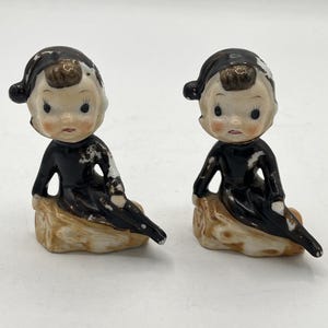 May include: Two vintage porcelain figurines of seated figures with black outfits and hats. The figures have rosy cheeks and are perched on tan-colored bases. The figurines have a distressed, antique appearance.