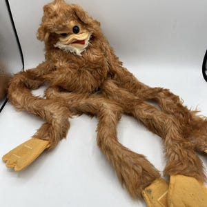 May include: A vintage brown plush toy with long, flexible limbs and a distinctive face. The toy features a duck-like beak, a black eye, and tan felt feet. The plush has a shaggy texture and is likely from the mid-20th century.