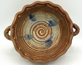May include: A round, hand-thrown ceramic bowl with a scalloped rim and two twisted side handles. The interior features concentric circular grooves in a warm brown clay tone, accented with three distinct blue painted brushstroke marks.
