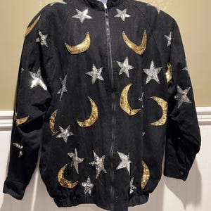 May include: Black jacket with a full zipper closure, adorned with silver star and gold crescent moon sequins. The jacket has a round collar and long sleeves. The jacket is displayed on a white mannequin.
