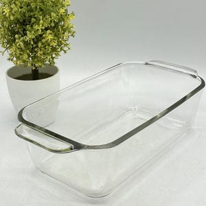 Puede incluir: Un molde rectangular de vidrio transparente para pan con asas integradas en ambos extremos cortos. El molde presenta paredes lisas y transparentes con una base plana, diseñado para hornear pan u otros alimentos en un horno estándar.