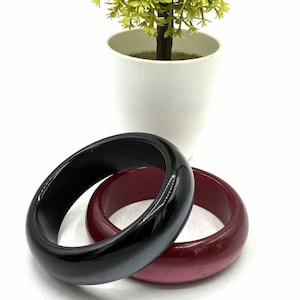 May include: Two smooth, thick bangle bracelets are positioned on a white surface. One bangle is a solid, glossy black, while the other is a deep, translucent burgundy red. Both bracelets feature a rounded, polished exterior surface and a circular shape.
