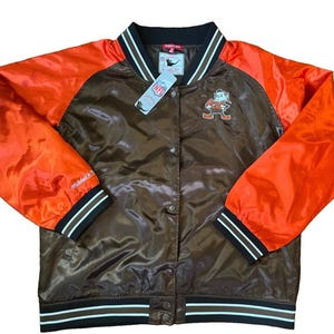 May include: A brown and orange satin varsity jacket featuring a snap-button front closure, striped ribbed collar, cuffs, and hem. The chest displays an embroidered Cleveland Browns logo, and an NFL tag is attached to the collar area.