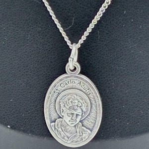 Puede incluir: Un collar con colgante ovalado de tono plateado que presenta un retrato en relieve de St. Carlo Acutis. El colgante cuelga de una cadena de tono plateado a juego. El texto St. Carlo Acutis está grabado en el borde superior del dije.