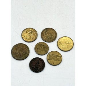 May include: A collection of seven circular metal arcade tokens in various shades of gold and bronze. The tokens feature embossed designs including a mouse character, the text Aladdin's Castle, Pavilion Amusements, and Discovery Zone.