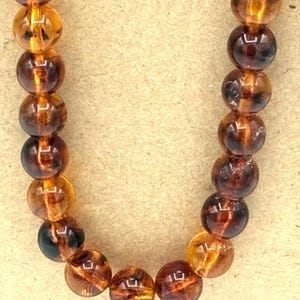 May include: A necklace composed of two strands of spherical beads in varying shades of translucent amber, ranging from light honey yellow to deep dark brown, featuring a polished, glossy finish that reflects light across the rounded surfaces.