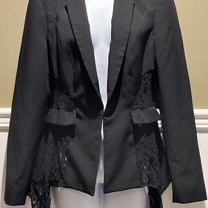 Könnte beinhalten: Ein schwarzer Blazer mit fallendem Revers und zwei Vordertaschen. Die Seiten des Blazers weisen schwarze Spitzeneinsätze auf. Der Blazer wird auf einer weißen Schaufensterpuppe präsentiert.