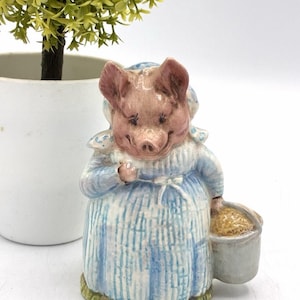 Può includere: Statuina in ceramica di un maiale sorridente con un vestito blu a righe bianche e un cappello. Il maiale tiene in mano un piccolo secchio pieno di una sostanza beige. La figura è dipinta con colori rosa, blu e beige.