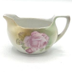 May include: A vintage porcelain creamer with a pink rose floral design. The creamer has a curved handle and a spout for easy pouring. The exterior features a gradient of colors, transitioning from gold to green, with a pink rose painted on the front.