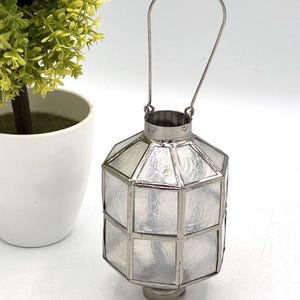 Puede incluir: Una linterna hexagonal construida con paneles de vidrio transparente texturizado unidos por un marco de metal de tono plateado. La linterna cuenta con una abertura cilíndrica en la parte superior y un asa de metal arqueada y delgada unida al marco, lo que permite colgarla o transportarla fácilmente.