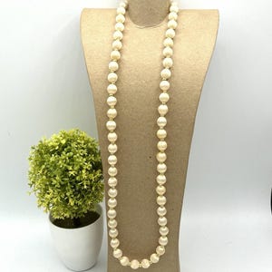May include: A long necklace featuring a single strand of round, cream-colored satin-finish beads alternating with small, polished gold-tone spacer beads. The necklace is displayed on a tall, neutral-toned, cylindrical jewelry display stand.