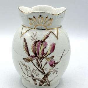 Pode incluir: Um vaso de cerâmica branca com corpo bulboso e borda ondulada. O vaso apresenta um design floral com íris roxas e marrons. Detalhes dourados estão presentes na borda e ao redor do design floral.
