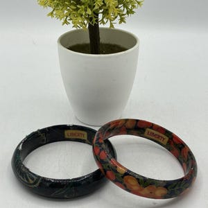 May include: Two wooden bangles with floral designs and the word "LIBERTY" printed on them. One bangle is black with a floral pattern, and the other is decorated with orange and red flowers on a black background. A small potted plant is in the background.