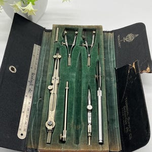 May include: An open black case with a green velvet interior holds several vintage drafting tools. Included are compasses, dividers, and a ruler. The case is labeled "ACADEMIC" and "TECHNICAL SUPPLY CO SCRANTON PA MADE IN GERMANY". A small plant is in the background.