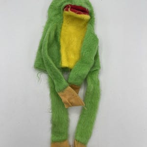 May include: A plush frog puppet with a green body, yellow chest, and tan feet. The frog has a red mouth, yellow eyes, and long, dangling legs. The puppet is made of a fuzzy material.