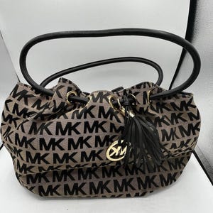 Op de afbeelding: Een beige en zwarte handtas met een trekkoordsluiting en een zwart handvat. De tas heeft een herhalend "MK" logo-patroon. Een goudkleurige bedel met het "MK" logo en een zwarte kwast hangen aan het trekkoord.