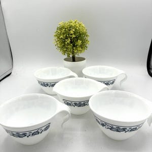 May include: A set of five white ceramic cups with a blue floral pattern around the exterior. Each cup features a wide, open bowl shape and a curved handle extending from the side. The cups are arranged in a group with a small green plant behind.