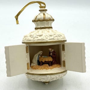 Könnte beinhalten: Ein cremefarbener, glockenförmiger Anhänger mit goldenen Akzenten und einem goldenen Aufhängeband. Der Anhänger verfügt über zwei aufklappbare Türen, die eine Miniatur-Krippenszene mit Figuren um eine Futterkrippe mit Heu im Inneren enthüllen.