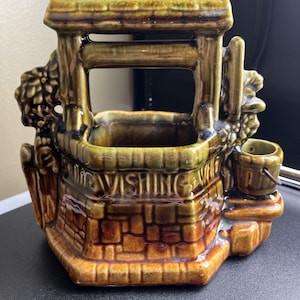 Pode incluir: Um vaso de cerâmica em forma de poço dos desejos com um design rústico. O poço apresenta um esquema de cores castanho e verde, com uma base semelhante a tijolos e um pequeno balde. As palavras "Wishing Well" são visíveis.