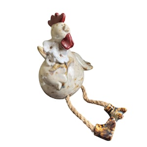 May include: A ceramic chicken figurine with a round, speckled cream-colored body, a red comb and wattle, and white floral accents. The figure features two dangling legs made of thick, natural-colored rope with brown and tan ceramic feet.