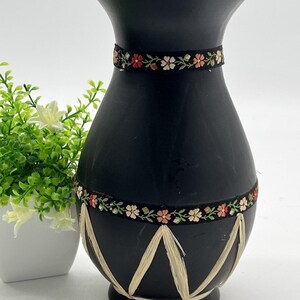 Könnte beinhalten: Eine schwarze Vase mit breitem Oberteil und dekorativem Blumenband um den Hals. Die Vase hat eine strukturierte Oberfläche und ist mit einem Band aus gewebtem Bast verziert. Die Vase ist etwa 25 cm hoch.