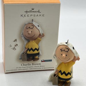 May include: A Hallmark Keepsake Halloween ornament featuring Charlie Brown in a ghost costume. The figurine has a yellow shirt with a black zig-zag pattern, brown shoes, and a small bag. The box is orange and white.