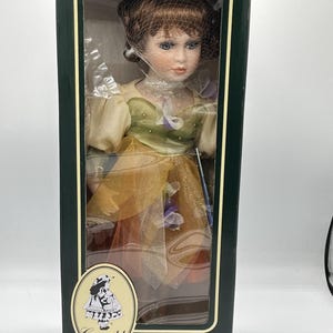 May include: A porcelain doll from the Geppeddo Fairy Tale Series, packaged in a green box. The doll has brown hair, blue eyes, and wears a dress with gold and green accents. The box features an illustration of a girl and the text "Fairy Tale Series."