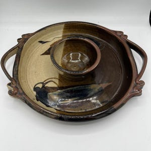 May include: A handmade ceramic serving platter with a built-in dip bowl. The oval platter has two handles and a two-tone design in brown and tan, with abstract black and blue accents. A decorative piece.