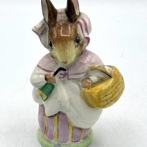 May include: A ceramic figurine of a rabbit wearing a pink and white bonnet and dress, holding a yellow basket and a green object. The rabbit has brown ears and a white face. The figurine is on a green base.