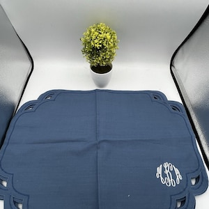 Puede incluir: Juego de manteles individuales azul marino con borde festoneado y monograma blanco. Una pequeña planta en maceta con follaje amarillo-verdoso se encuentra en el fondo. Los manteles individuales son de un material suave, posiblemente lino.