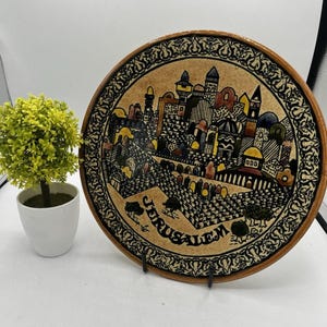 May include: A decorative ceramic plate featuring a detailed illustration of Jerusalem. The plate has a brown border and a beige center with colorful buildings and the word "JERUSALEM". The plate is displayed on a black stand.